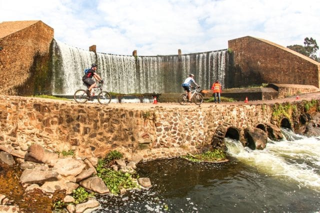 Riders pass below the waterfall cascading over the weir of Lake Heritage during Stage 2 of the Glacier Cradle Traverse, on Saturday the 6th of May 2017. Photo by Oakpics.com.